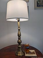 Light bronze minimalist table lamp with white fabric drum shade placed on a round wooden table, showing the full height and scale with a soda can for size reference.