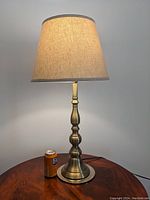 Photo shows a light bronze metal table lamp with a beige linen shade placed on a wooden surface. The electrical cord is visible behind the lamp.