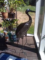 Photo of a black metal crane statue standing on thin metal legs on a porch floor, next to gardening supplies and plants, showing scale and condition.