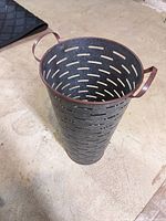 Top-down and angled views of a tall cylindrical metal container with horizontal oval perforations and two handles at the rim.