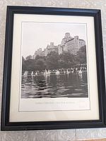 Framed black and white photo showing the Model Boat Pond with model sailboats and city buildings in the background, matted and encased in black frame.