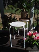 Two white metal round stacking tables shown outdoors on porch, one nested inside the other, surrounded by plants and flowers