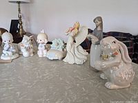 The lot of porcelain Easter themed figurines arranged on a table shows eight figurines including children, rabbits, an angel with wings, and a small car decoration.