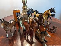 Group of porcelain and metal horse figurines on a wooden table, various colors and sizes visible, with a Fanta can and lamp in background