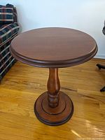 Round wooden Mid Century Modern table with central pedestal base and smooth finish, placed on wooden floor near plaid sofa and office chair.