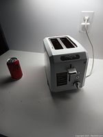 White Waring toaster on white surface next to a soda can for scale, plugged into wall outlet.