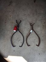 Two antique metal ice tongs on a concrete floor, one with a soda can placed in the claw for scale, showing their large size and vintage condition.