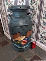 Front left angled view of vintage metal milk can with eagle design and removable lid, placed on stone floor with floral wallpaper background. Coke can for size reference.