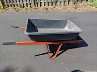 Side angle showing the plastic bed, orange frame, and black handles of wheelbarrow on paved surface