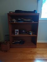 Wood bookcase with three shelves, medium brown finish, placed against a wall near a window. Some miscellaneous items on shelves not included in sale.