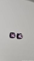 Two natural purple amethyst gemstones, cushion checkerboard cut, shown on a light background.