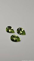 Three pear cut apple green peridot gemstones arranged on a white background, showing facet reflections and clarity.