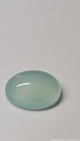 Oval cabochon Arctic Blue Chalcedony gemstone on white background showing translucent pale blue color and smooth polished surface.