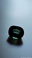 Photo of a single oval cut natural sapphire gemstone displaying teal green hue on a light background, showing its transparency and faceted surface.