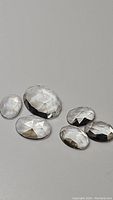 Six natural crystal quartz gemstones, transparent with fancy rose cut, varying oval/round shapes, smooth polished surface, displayed on a flat light grey surface.