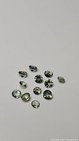 Group of 13 natural green apatite gemstones in various cuts and shapes on a plain background, showing their clarity and color.