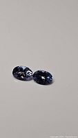 Pair of oval cut blue-purple tanzanite gemstones on a neutral background showing their size, color, and clarity.