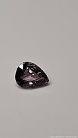 Close-up image of a pear cut natural purple spinel gemstone. The gem is well-faceted with a deep purple hue and measures approximately 7x5.2 mm.