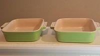 Two Le Creuset small square baking dishes side by side with green exterior and cream interior.