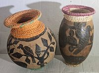 Pair of hand carved folk art vases with woven tops, displaying the full body with animal and leaf designs.