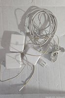 Four Apple MacBook chargers with cables coiled and intertwined, laid on a white surface. Some chargers include detachable AC prongs.