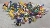 Top view showing an assorted collection of about 70 small Pokemon figures scattered on a surface. Multiple species and poses visible including Pikachu and Gengar.