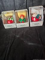 Photo showing three Noel ornaments in clear packages with 'Noel' branding on box, including Santa and elf figures.