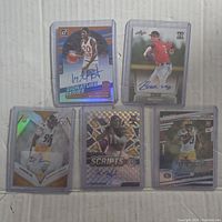 Photo showing five sports autographed cards in protective holders including basketball, baseball, and football cards with holographic and foil designs.