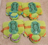 Top view showing all four TMNT Dream Lites plush night lights laid flat with green fuzzy bodies and colored masks in blue, orange, purple, and red. Each has the translucent light projector on the back with star patterns and turtle face.