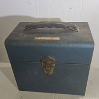 Blue metal carrying case with wear, viewed from front showing latch and handle on top.