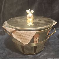 Brass warming dish with brass lid with leaf-shaped knob on top, brown paper inside for protection.