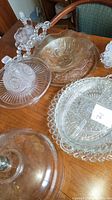A top-down view of various cut glass plates and bowls arranged on a wooden table, showing a range of clear and amber tinted glass items with cut patterns and a lidded glass cake stand.