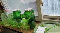 Green depression glass jars, drinking glasses, and dessert bowls on a wooden surface by window