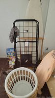 Ironing board, black metal rolling laundry cart, and white plastic woven laundry basket shown against basement wall. Additional items in background include a wooden folding chair, boxed laundry supplies, and other household items.
