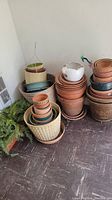 Photo showing various stacked and nested outdoor planters and pots including terracotta, ceramic, and plastic types, arranged in the corner of a room on a tile floor.