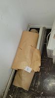 Two large rolls of rolled brown packing paper placed at corner of room near vent and heater.