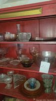 Red shelving unit with multiple glassware items including pink depression glass bowls, dishes, vases, and clear glass pieces.