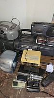 Full view of collection showing several radios, boomboxes, cassette players, and miscellaneous electronic devices on a wooden floor against a white wall