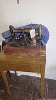 Singer sewing machine on wooden base atop sewing table, showing machine body, handwheel, foot pedal, and surrounding cords.