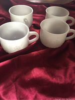 Four opaque white milk glass punch bowl cups with raised grape cluster fruit design on the side placed on a red satin fabric background.