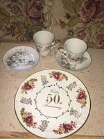 View of two tea cups, their matching saucers, and a large commemorative 50th anniversary plate displaying floral motifs and anniversary text.