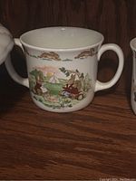 White ceramic child's mug with two handles and colorful Bunnykins rabbit scene around the sides standing on a wooden surface.