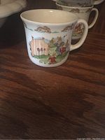 White ceramic children's mug with colorful Bunnykins bunnies playing with pulley device and fence, with running bunny border near rim, on wooden surface