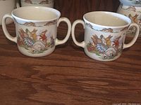 Front view showing two white ceramic mugs with double handles and colorful Bunnykins rabbit illustrations in countryside scenes.