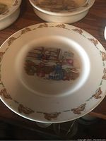 Front side of Bunnykins plate showing the detailed illustration of rabbits in a shop and a border pattern of rabbits.