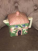 Front and side view of a ceramic tea pot shaped like a country cottage showing detailed paint and design work with a pink tiled roof lid.