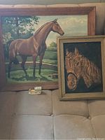 Two framed horse artworks resting on a surface: a larger horse landscape painting and a smaller textured horse head artwork.