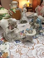 Porcelain Victorian couple seated at small tea table with tea set including teapot, cups, and pastry plate on lace tablecloth.