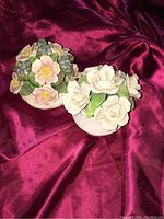 Two porcelain flower arrangements on dark red fabric; one with pink and blue flowers, the other with white roses.