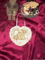 Three teddy bear collectibles including stuffed toy, heart-shaped plaque, and formal teddy bear figurine on red fabric.